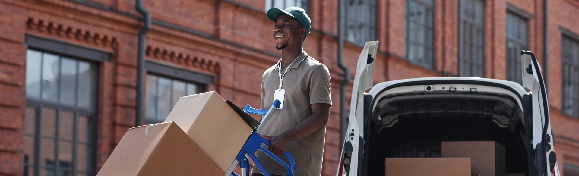 Lieferbote transportiert Kartons mit einer Sackkarre aus einem Lieferwagen vor einem Backsteingebäude.